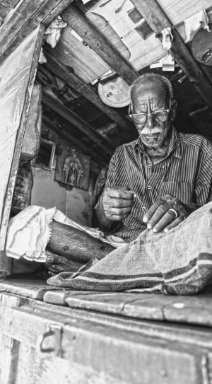 grayscale photography of man wearing eyeglasses while sewing clothe thumbnail
