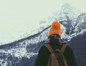 photo of person wearing orange knit hat facing through mountain filled with pine trees thumbnail