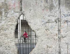 black haired female riding on bicycle seen through hole in gray concrete wall thumbnail