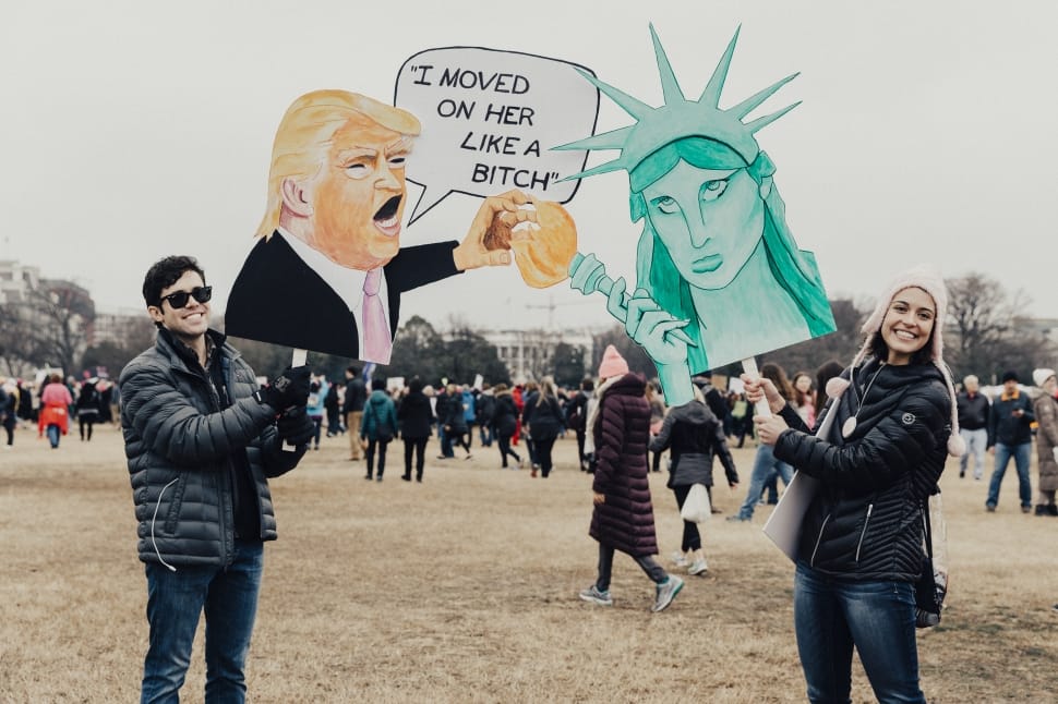 man and woman holding cutout of donald trump and statue of liberty during daytime preview