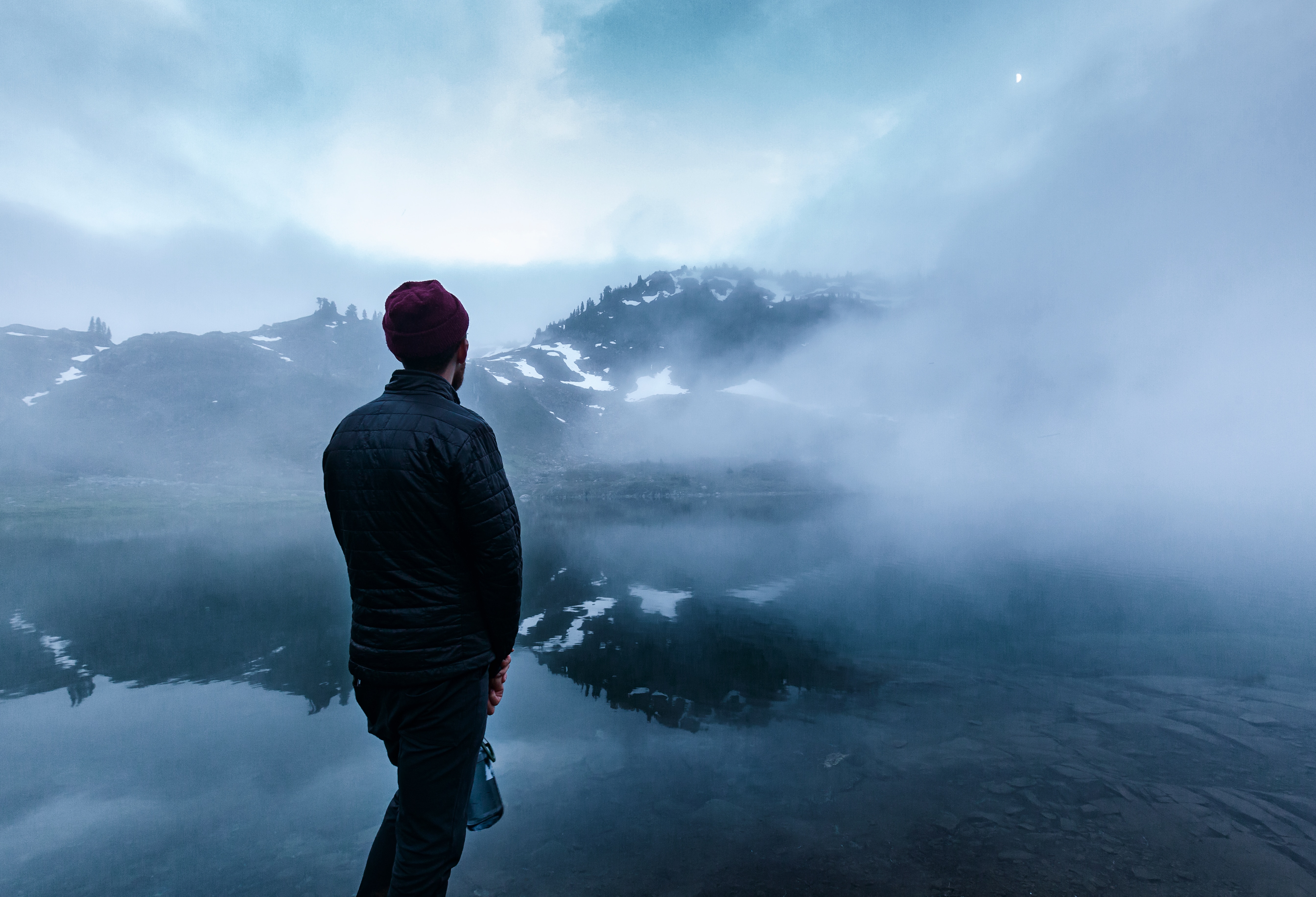 man wearing bubble jacket standing in front of body of water with fog