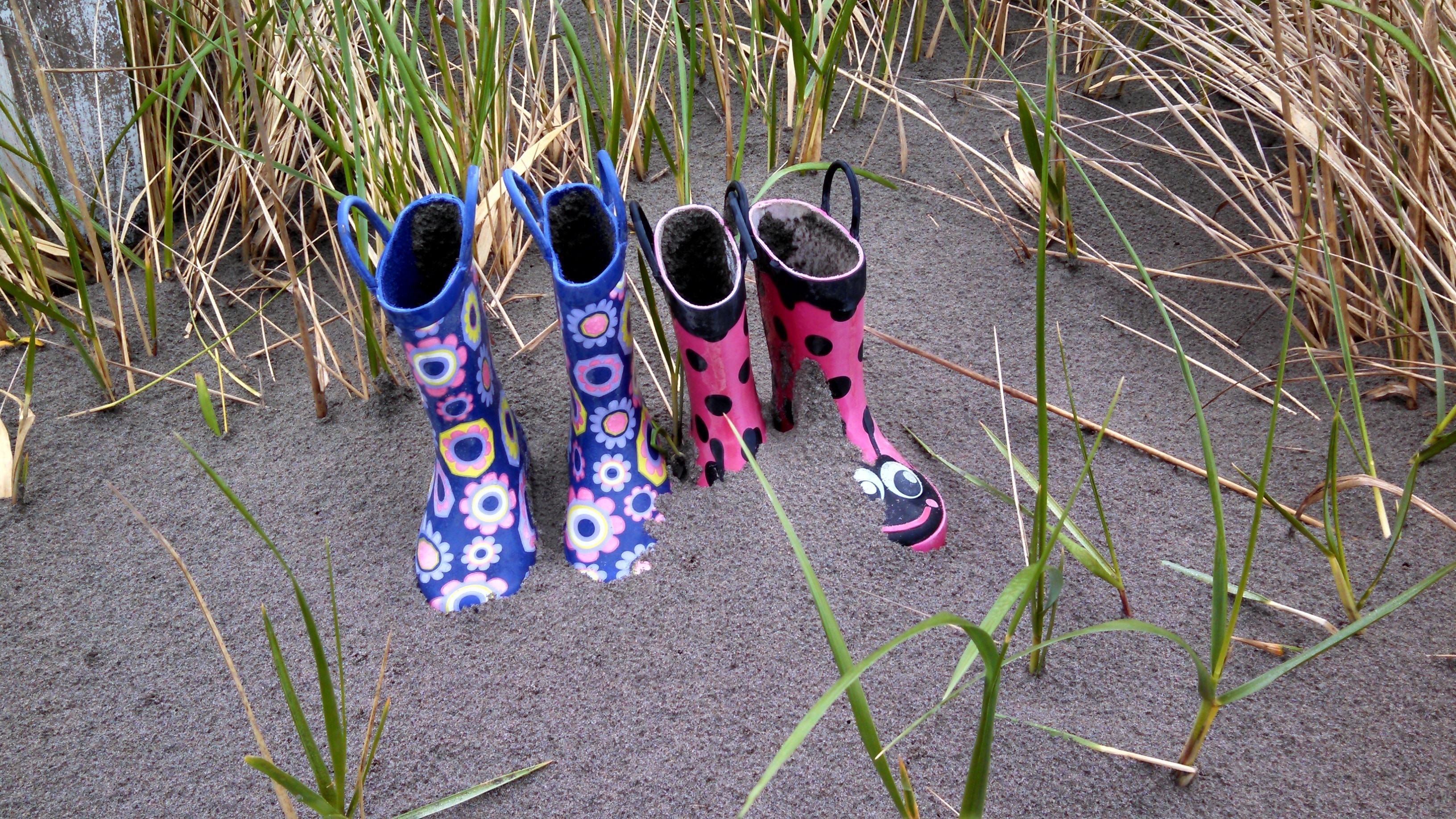 two pair of rainboots on sand surrounded by grasses