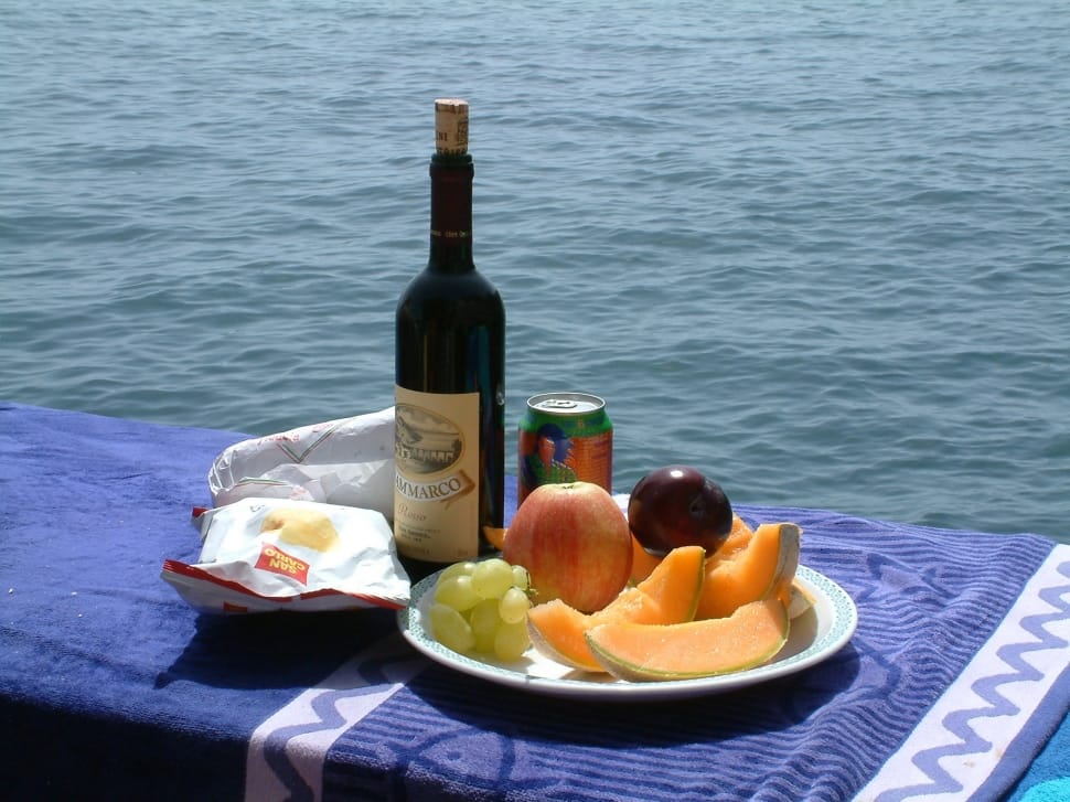 beige labelled wine bottle beside green can, white plastic pack and round white plastic plate with assorted fruits placed on blue and white textile near body of water during daytime preview