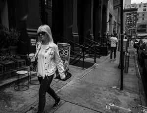 gray scale photo of woman walking on sidewalk near building thumbnail