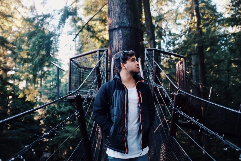 man in black zip-up jacket and white inner shirt on hanging bridge and tree bark view behind preview