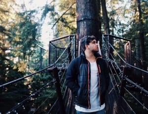 man in black zip-up jacket and white inner shirt on hanging bridge and tree bark view behind thumbnail