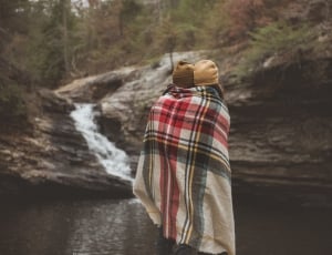 two person embracing while covering plaid blanket beside body of water thumbnail