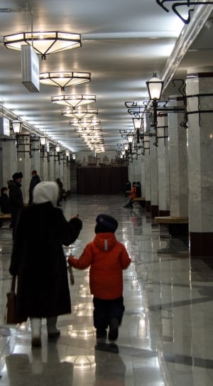 woman in black over coat with toddler in red jacket walking on white floor tiles thumbnail