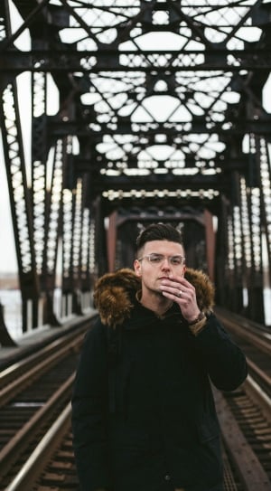 man in black parka coat with eyeglasses and ring on railroad bridge during daytime thumbnail