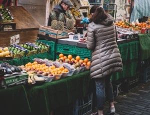 black haired woman wearing grey jacket and blue jeans standing on grey concrete crick road near assorted fruits stand thumbnail