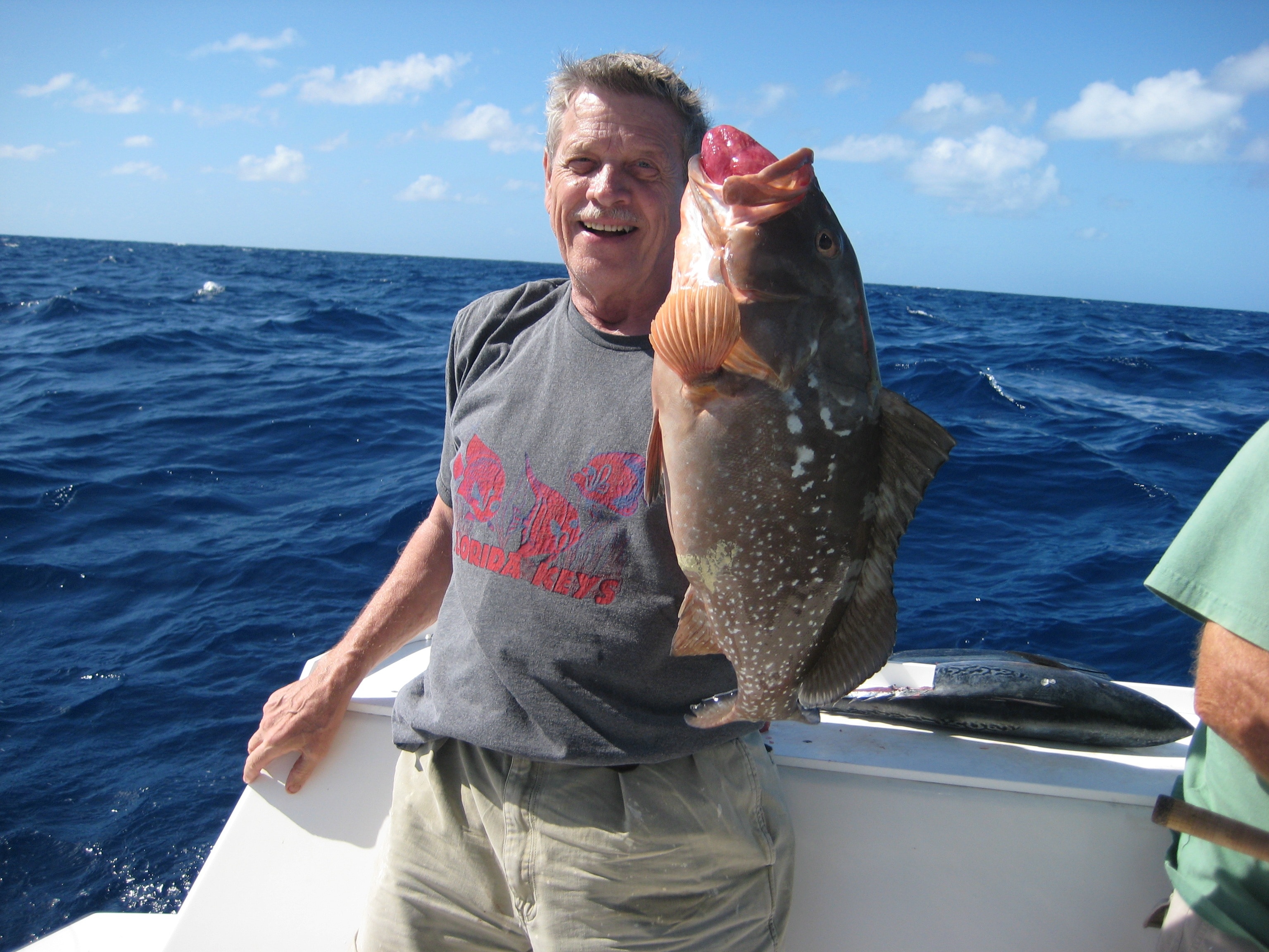 man in gray shirt and white pants holding gray fish standing on motorboat under blue and white sky during daytime