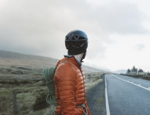man wearing orange bubble jacket facing grey road and clouds view thumbnail