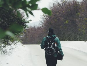 person in green jacket, black pants and black and gray backpack walking on snow covered road surrounded with trees under white sky during daytime thumbnail