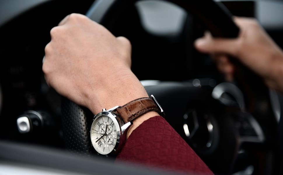 person wearing red long sleeve shirt and round silver chronograph watch with brown leather strap holding car steering wheel preview