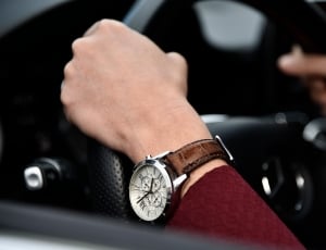 person wearing red long sleeve shirt and round silver chronograph watch with brown leather strap holding car steering wheel thumbnail