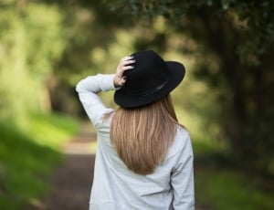 woman wearing white dress shirt and black fedora hat while holder her hat near green tree during daytime thumbnail