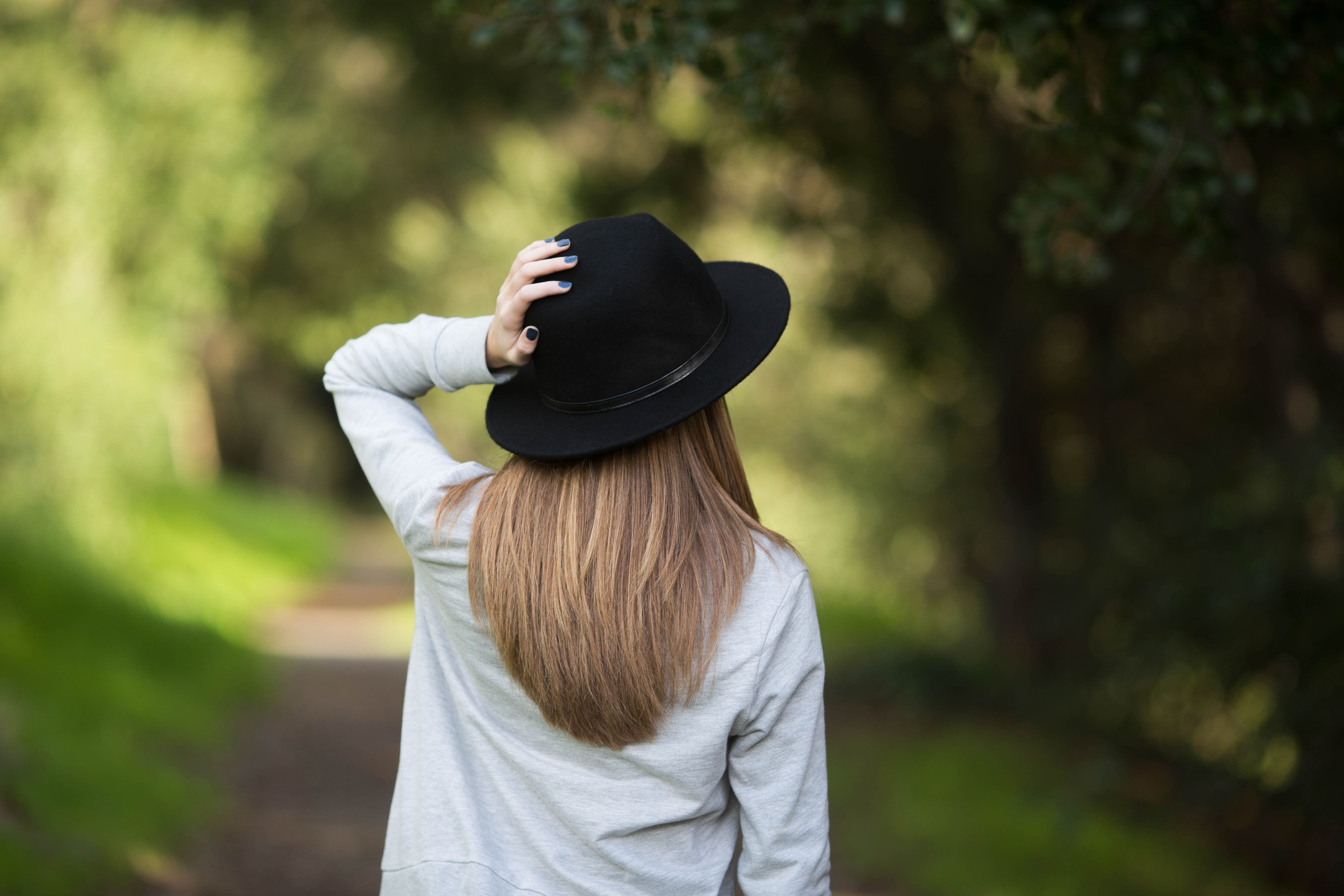 woman wearing white dress shirt and black fedora hat while holder her hat near green tree during daytime