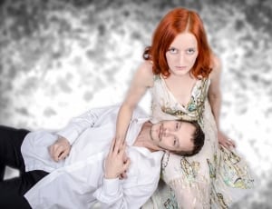 man in white dress shirt lying on woman in white,green,brown and yellow plunging neckline sleeveless dress thumbnail