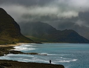 silhouette of man fishing in front of ocean beside green mountain during cloudy day thumbnail