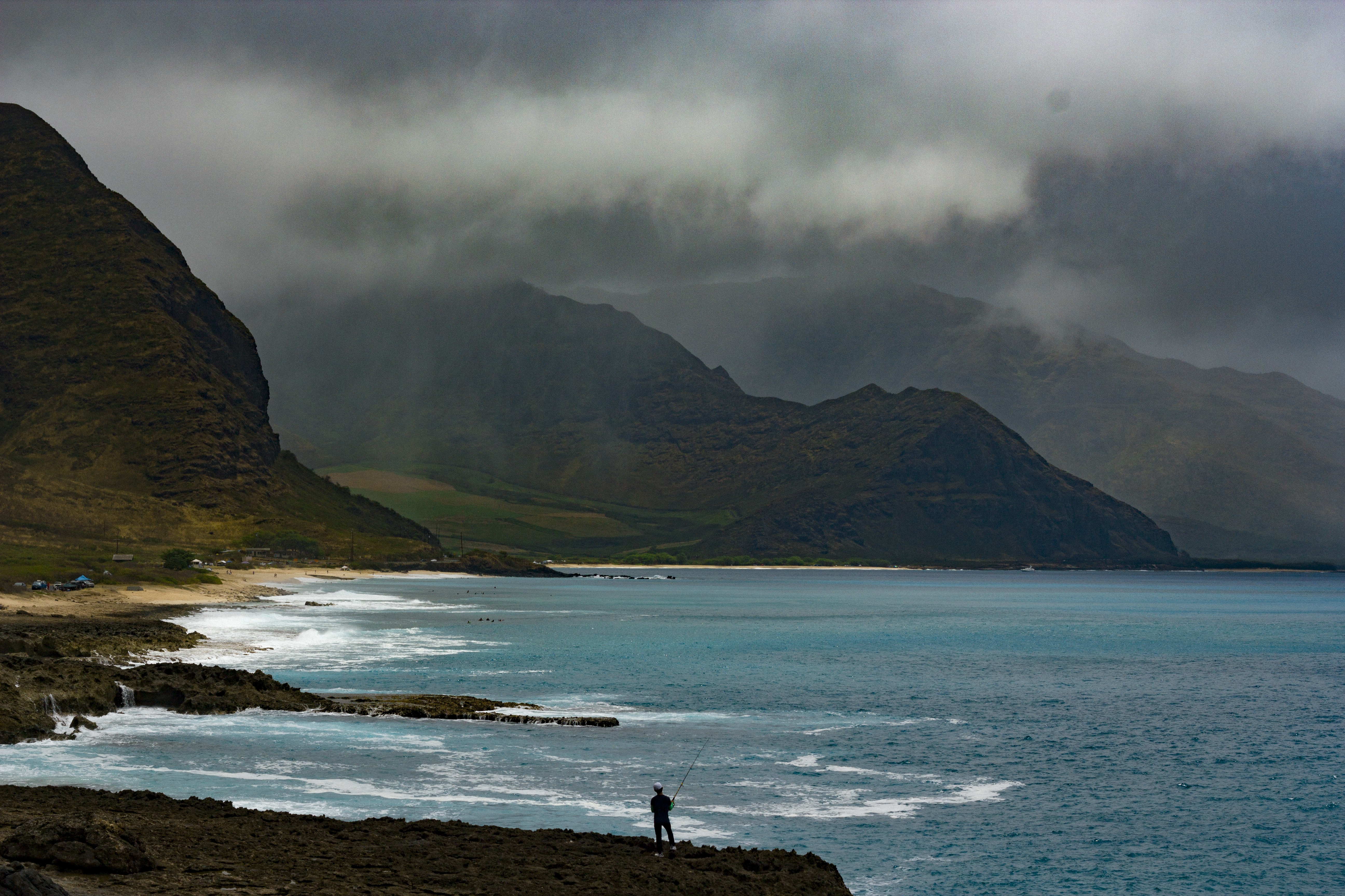 silhouette of man fishing in front of ocean beside green mountain during cloudy day