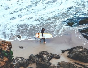 man in black surfing suit holding white wake board while walking near black rocks during daytime thumbnail