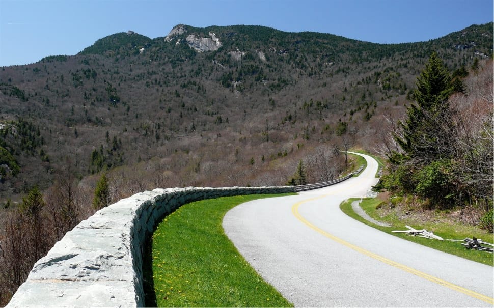 grey concrete road heading to mountain beside cliff with grey concrete barrier and trees under clear sky during daytime preview