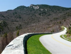 grey concrete road heading to mountain beside cliff with grey concrete barrier and trees under clear sky during daytime thumbnail