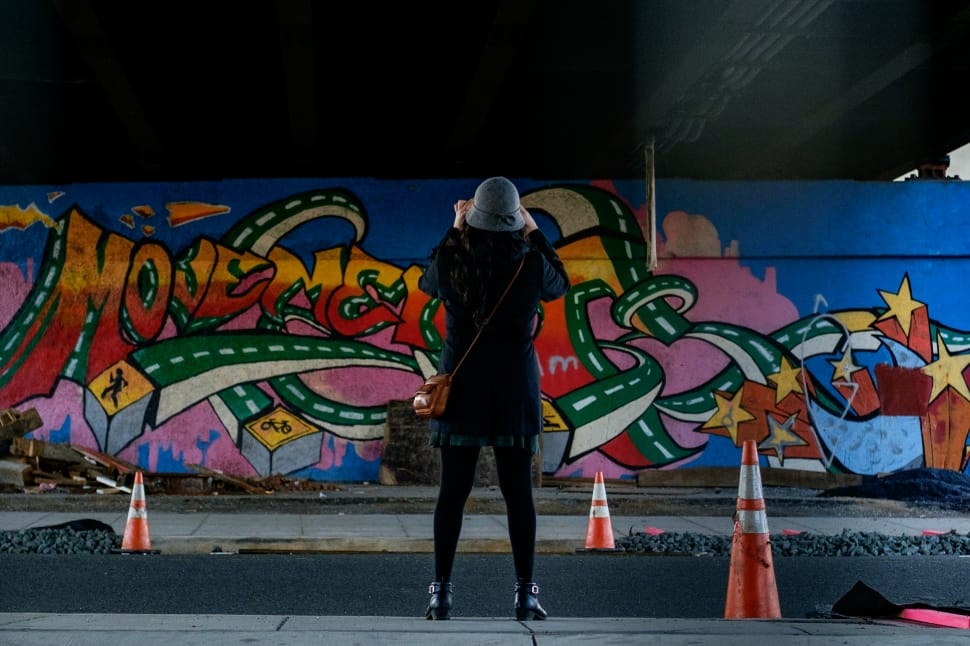 person wearing a blue hat taking picture of wall grafitti during daytime preview