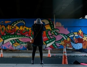 person wearing a blue hat taking picture of wall grafitti during daytime thumbnail
