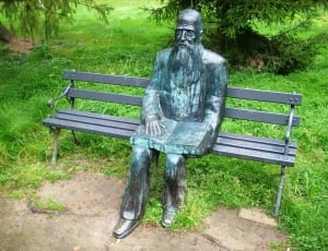man sitting on bench with open book grey and green statue thumbnail