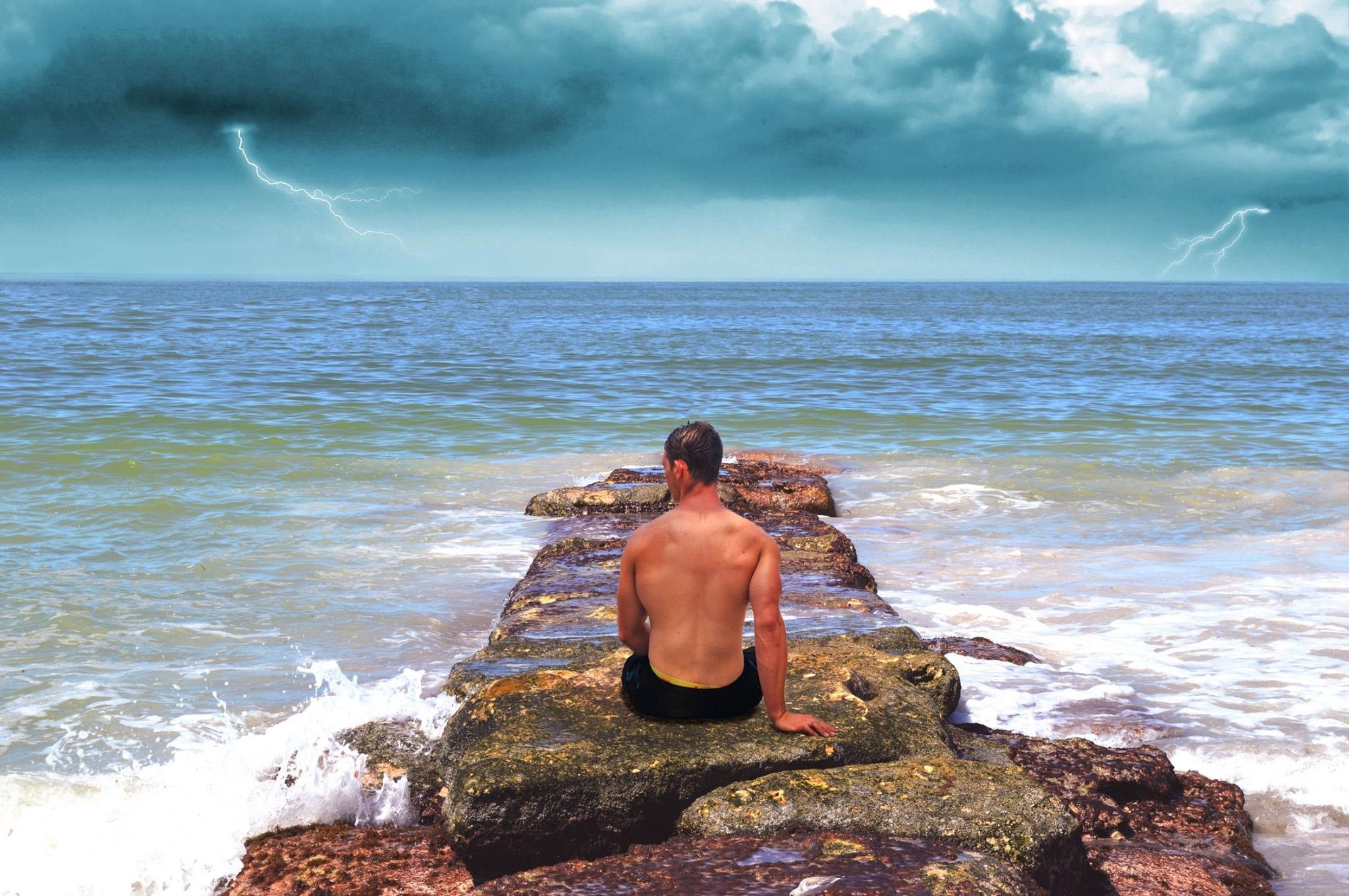 male in black shorts sitting on rock near beach