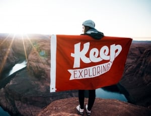 woman holding white and red keep exploring flag on top of mountain during daytime thumbnail