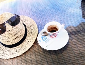 ceramic mug and saucer filled with coffee and brown hat and dark lens sunglasses thumbnail