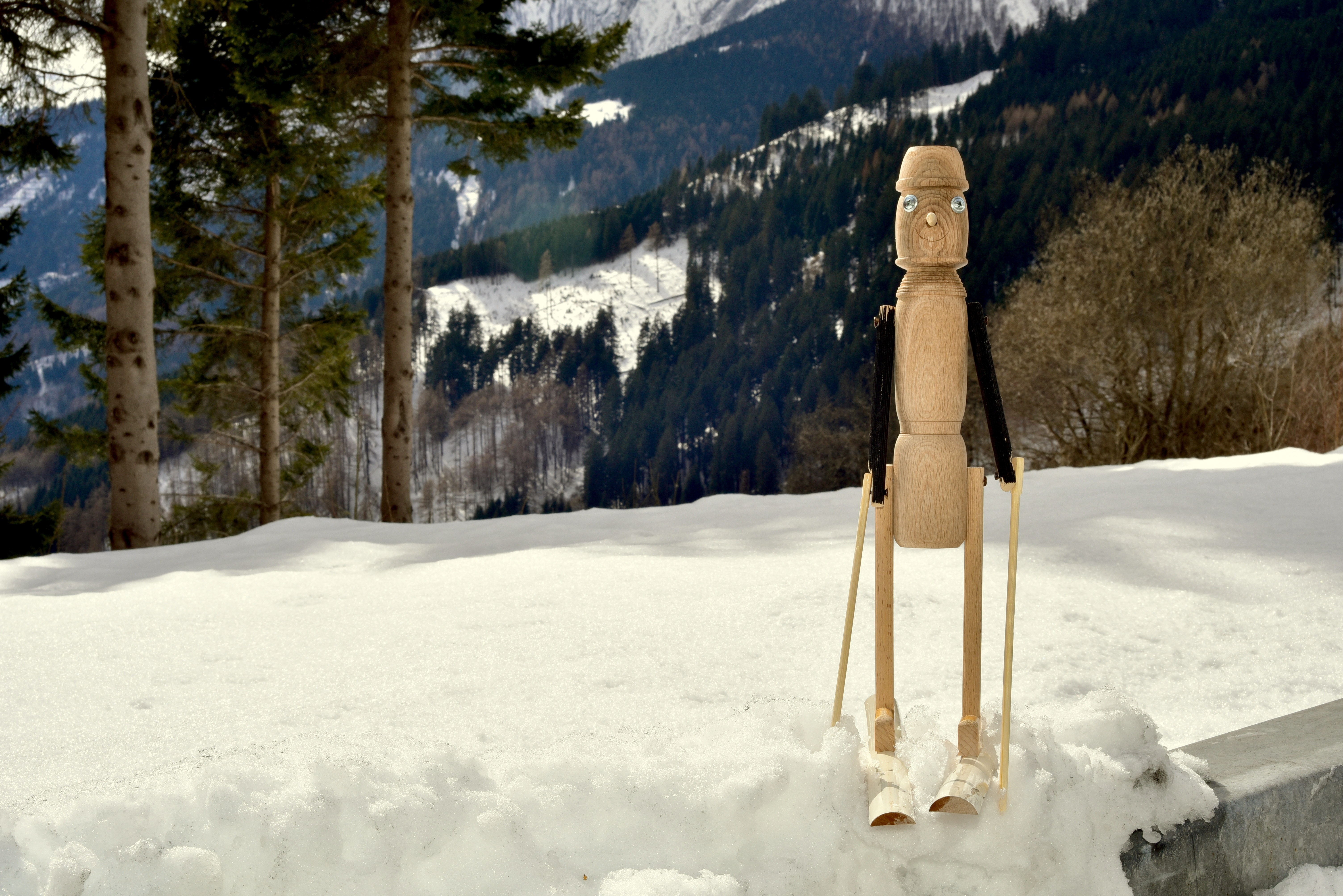 brown and black wooden puppet standing on snow surrounded with trees at daytime