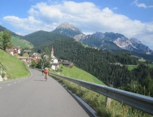 two person riding bikes in the middle of the road beside railings with view of town and mountains under blue sky and white clouds thumbnail
