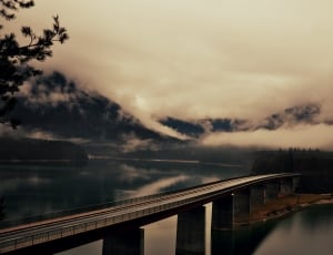 black and gray bridge over body of water across mountain covered with white clouds thumbnail