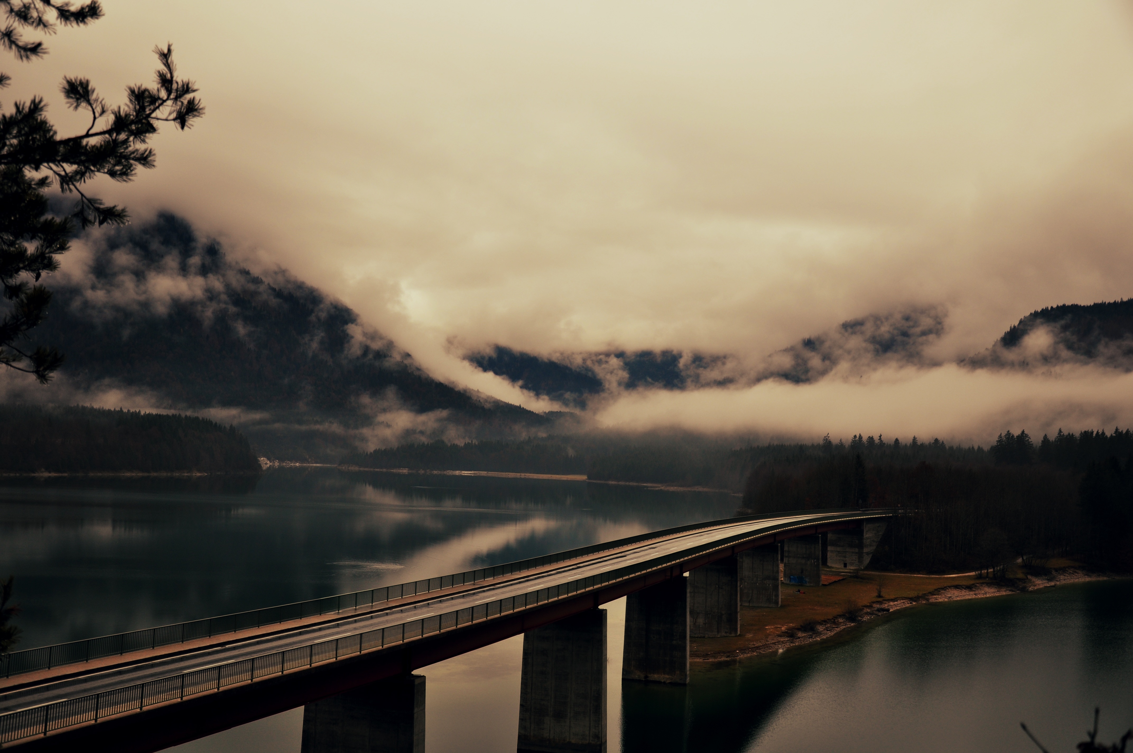 black and gray bridge over body of water across mountain covered with white clouds