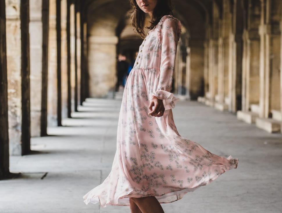 woman in pink long-sleeved tea-length dress standing in between concrete columns during daytime preview