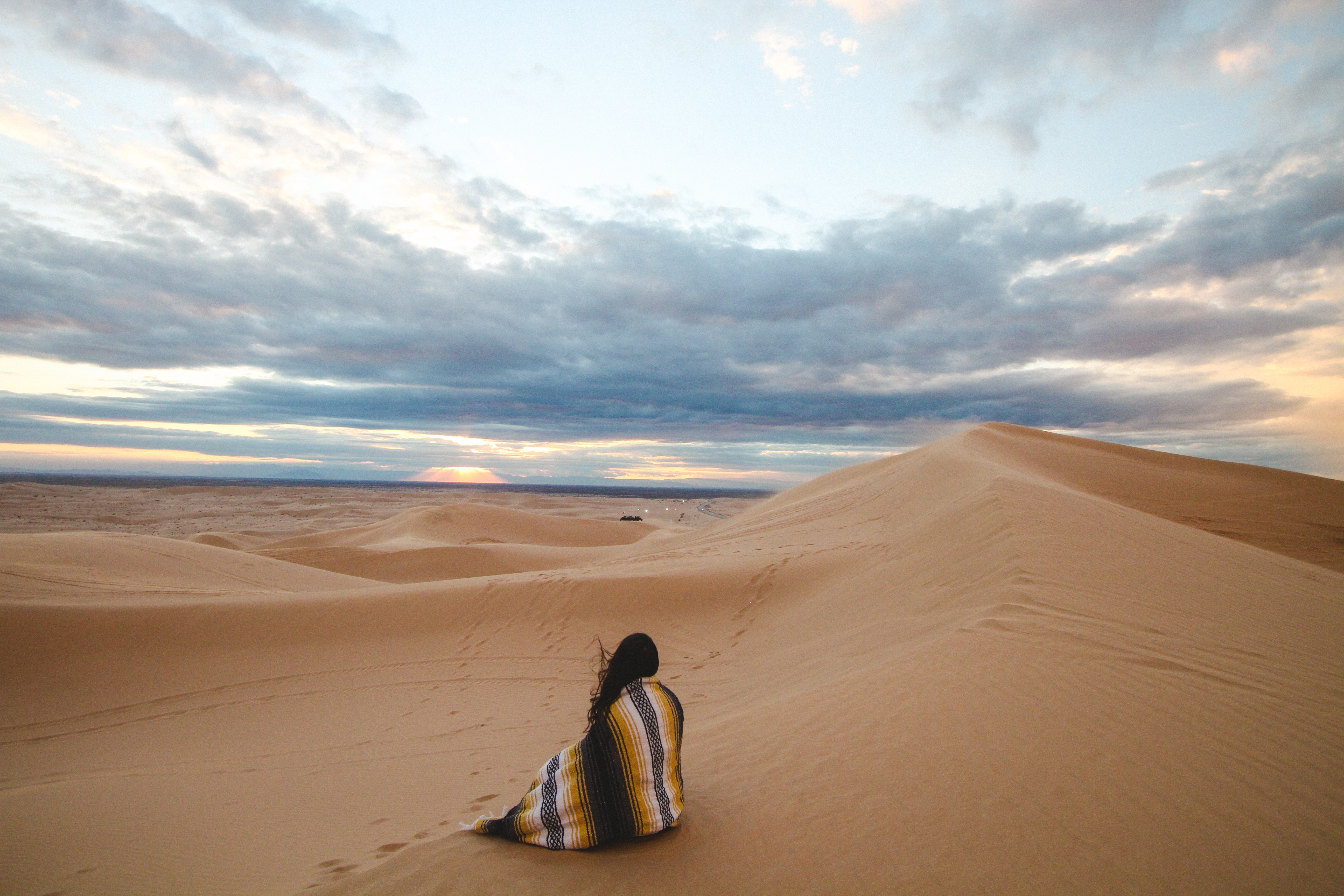 woman covered with brown and blue stripe blanket sitting on dessert under white clouds