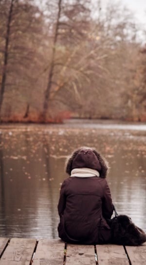 selective focus photo of person with brown parka jacket sitting beside the body of water thumbnail
