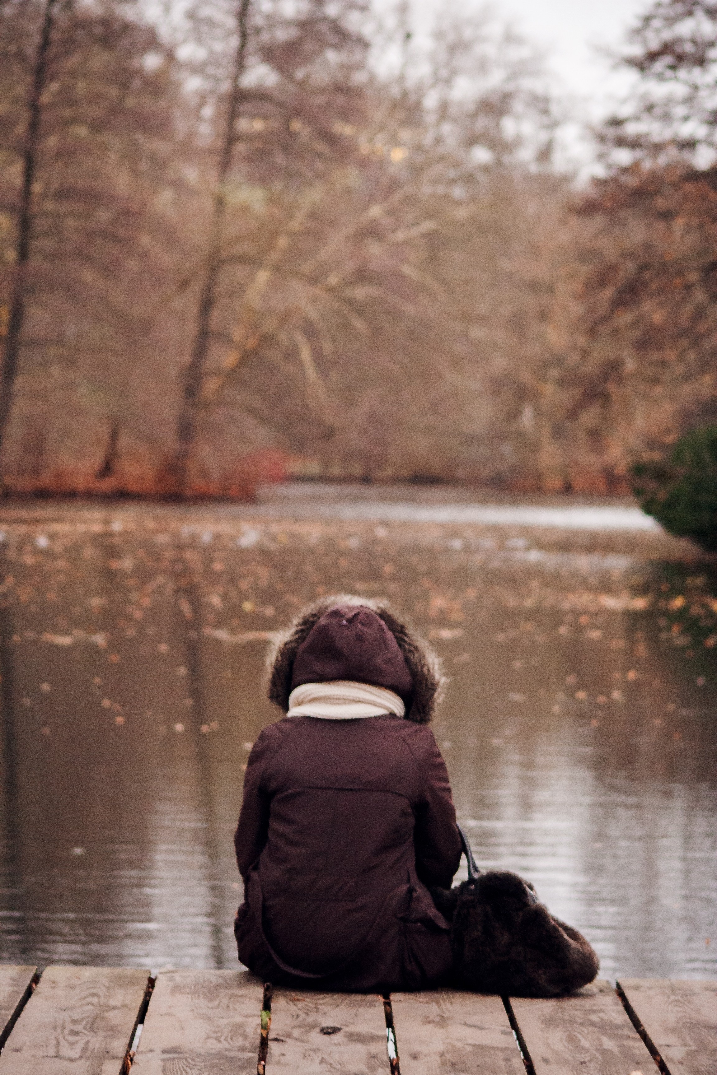 selective focus photo of person with brown parka jacket sitting beside the body of water
