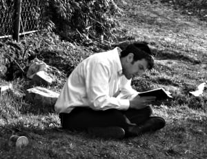 grayscale photo of man reading a book while sitting on grass thumbnail