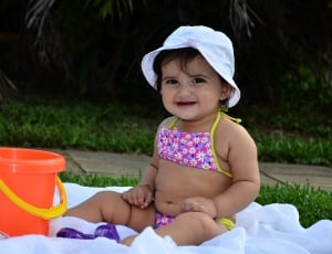 girl in pink, purple, and white floral string bikini and white hat sitting on white textile in front of orange plastic bucket thumbnail