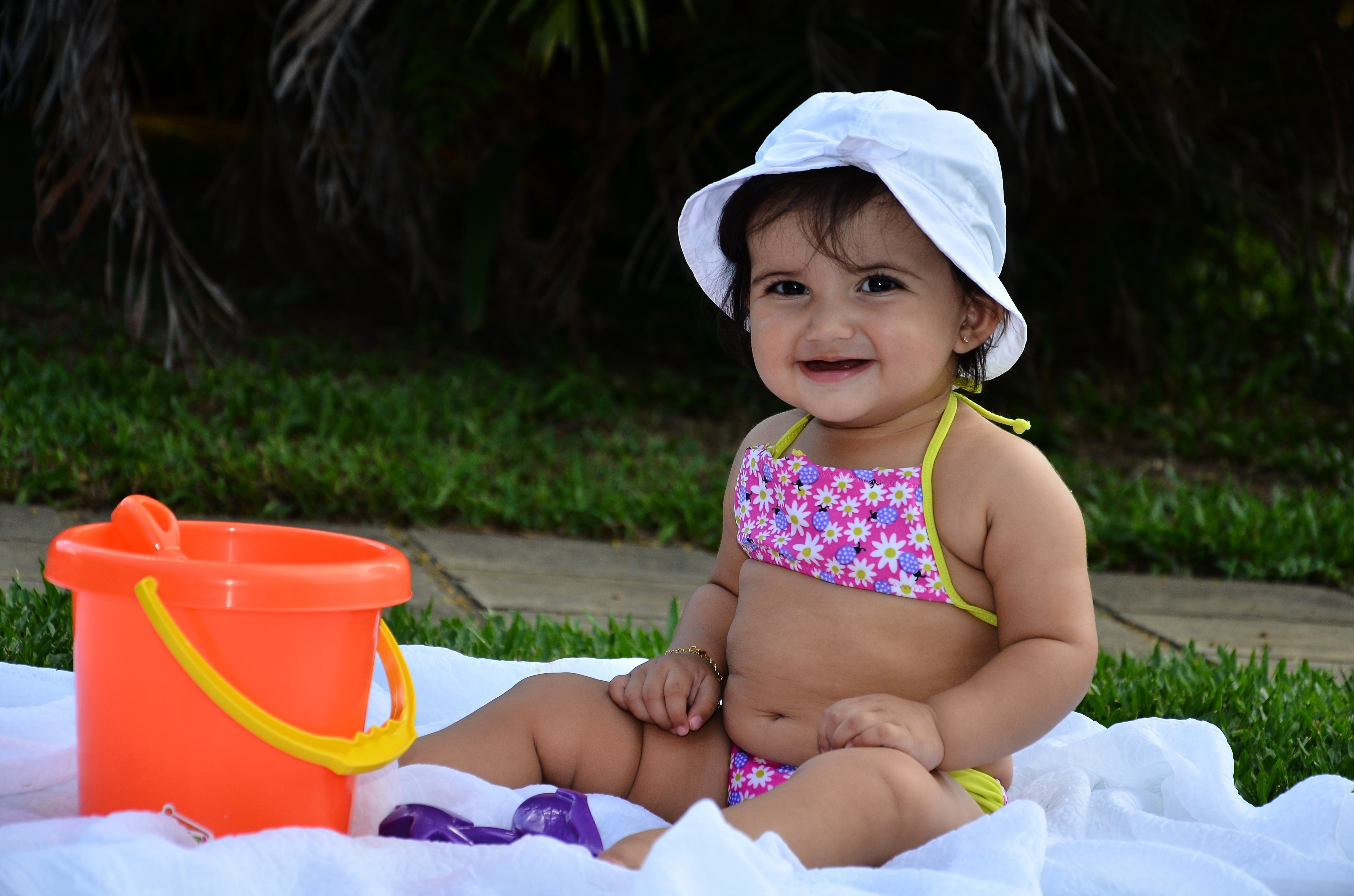 girl in pink, purple, and white floral string bikini and white hat sitting on white textile in front of orange plastic bucket