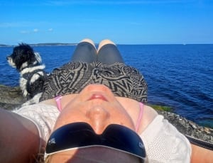 photo of woman in gray framed black sunglasses, white top, pink strap bikini top, gray capri pants with gray and black textile near medium black and white long coated dog near ocean thumbnail