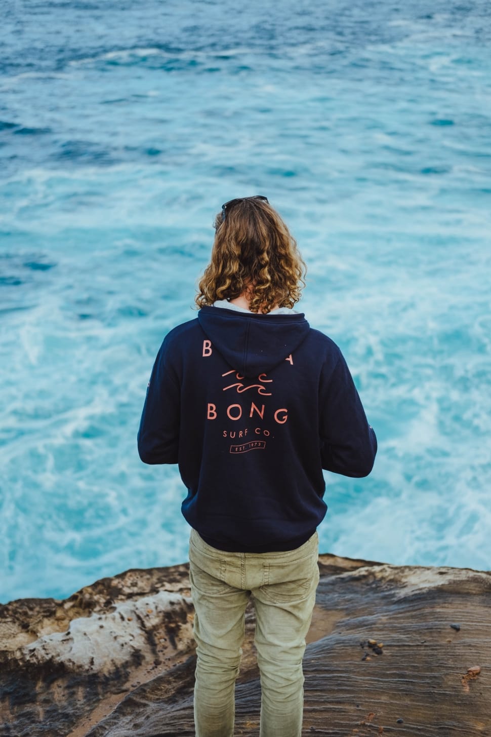 person with black printed hoodie with khaki pants on rock clip near on open body of water preview