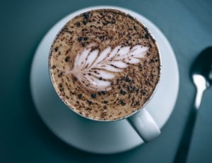 selective focus of creamy latte on white teacup on saucer near stainless steel teaspoon thumbnail