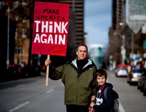 man in gray jacket holding a sign make america think again thumbnail