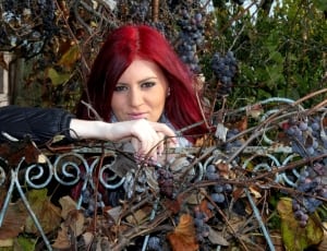 red haired woman leaning on steel fence with grape vines thumbnail