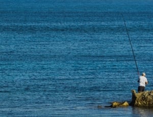 man holding fishing rod standing on stone near sea photo thumbnail